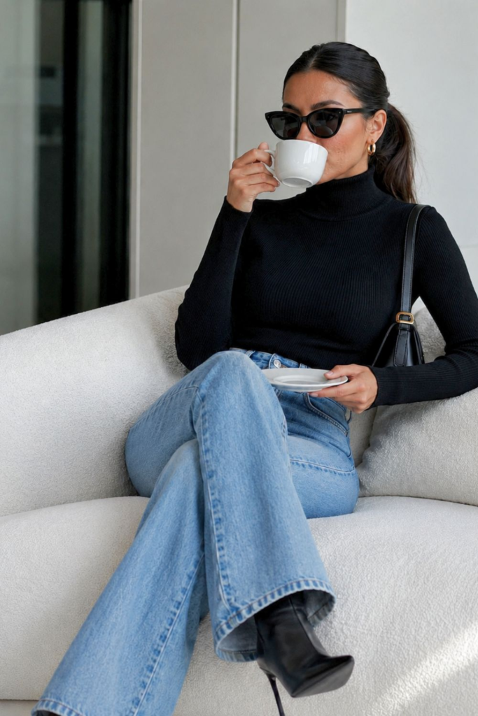 Woman wearing a black ribbed turtleneck and light blue high-waisted flared jeans with black pointed ankle boots, sitting on a cream sofa and drinking coffee in a minimalist modern interior
