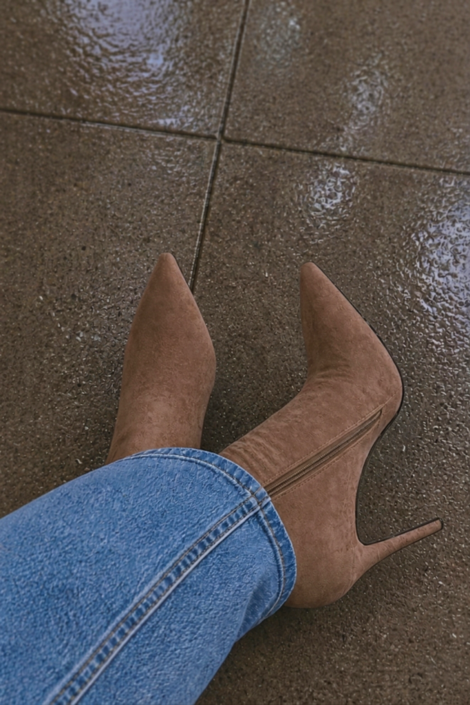 Close-up of a person wearing stylish tan suede boots in the rain, featuring a high heel and pointed toe, with light wash denim jeans and wet pavement reflections.