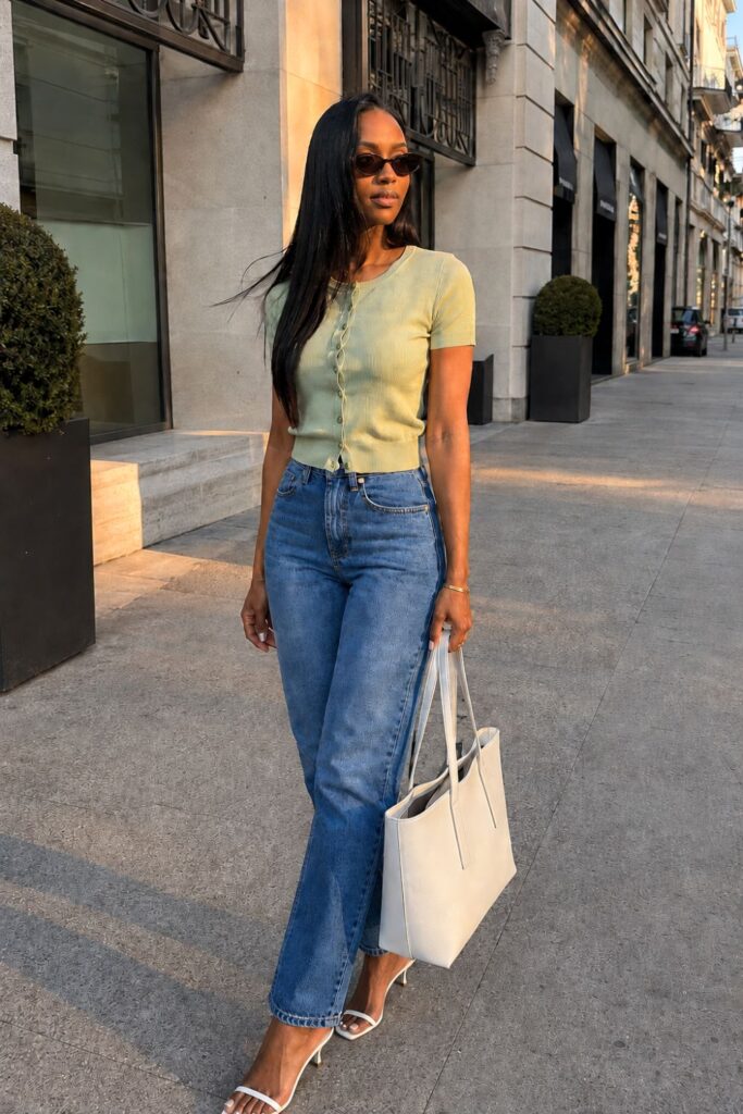 Black woman wearing a sage green cardigan, high-waisted blue jeans, white strappy heels, and narrow tortoiseshell sunglasses on a luxury city street during golden hour