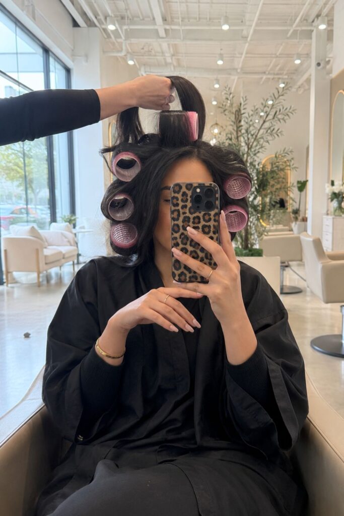 Woman with voluminous blowout in pink rollers at a luxury salon, showing healthy, glossy hair for an expensive look on a budget