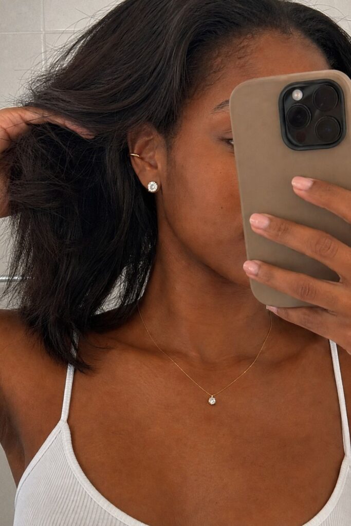 Woman wearing minimalist gold accessories, including a delicate pendant necklace, stud earrings, and ear cuff, paired with a white tank top
