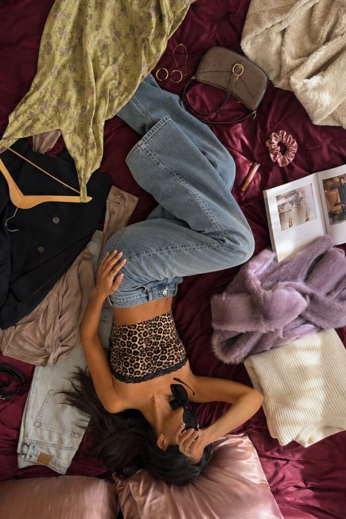 Young woman discovering her personal style while planning outfits on a burgundy bed surrounded by a capsule wardrobe, fashion accessories, and wide-leg jeans