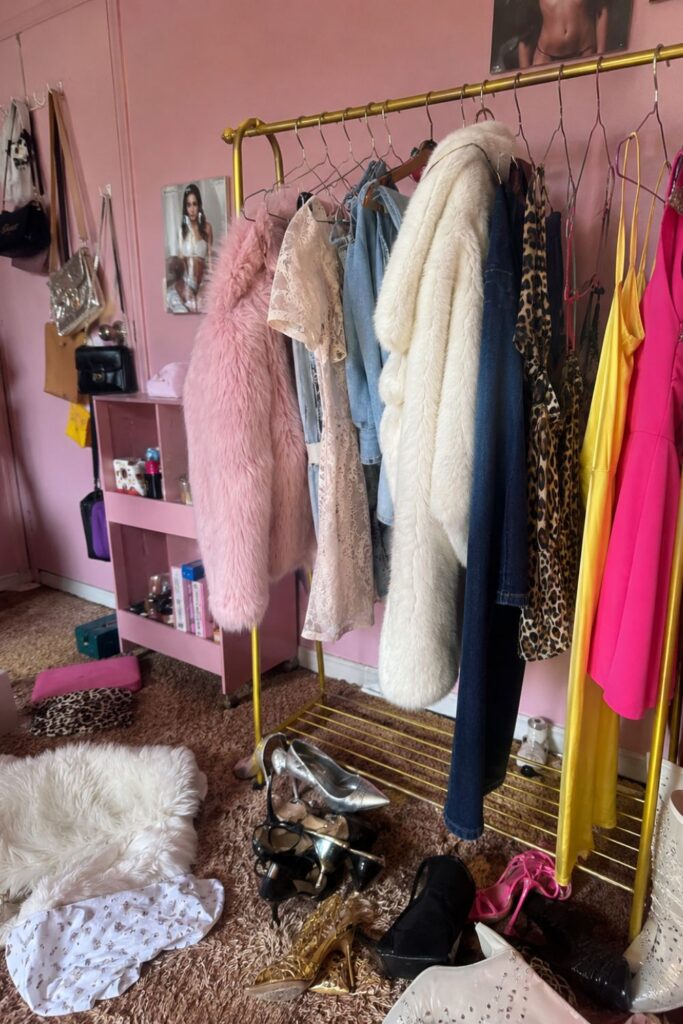 Colorful dressing room with a clothing rack full of Y2K and vintage-inspired outfits, faux fur coats, dresses, denim, and scattered heels, illustrating the process of decluttering a closet to define your personal style