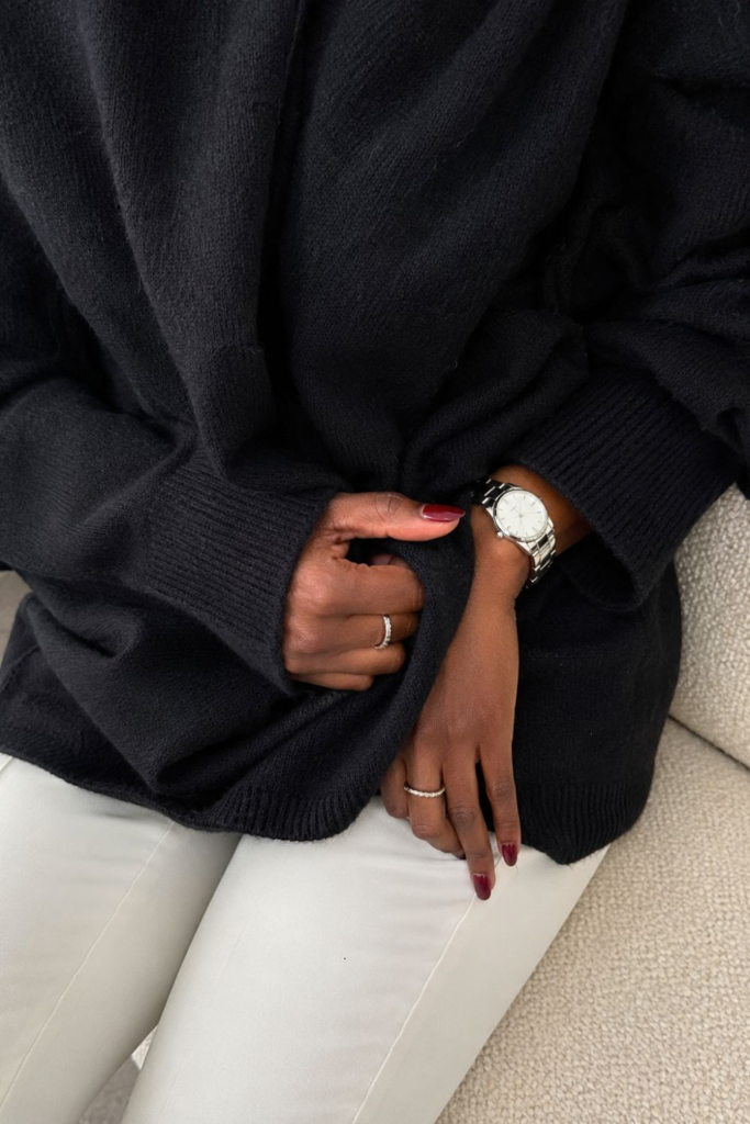 Close-up of elegant accessories featuring a silver watch, stacked rings and burgundy nails paired with an oversized black knit sweater and white trousers