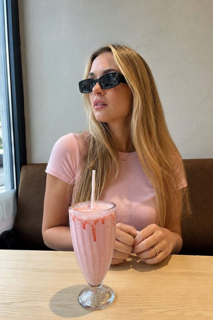 Fashion accessories featuring black rectangular sunglasses, a delicate gold necklace and ring styled with a light pink top while enjoying a strawberry milkshake at a café