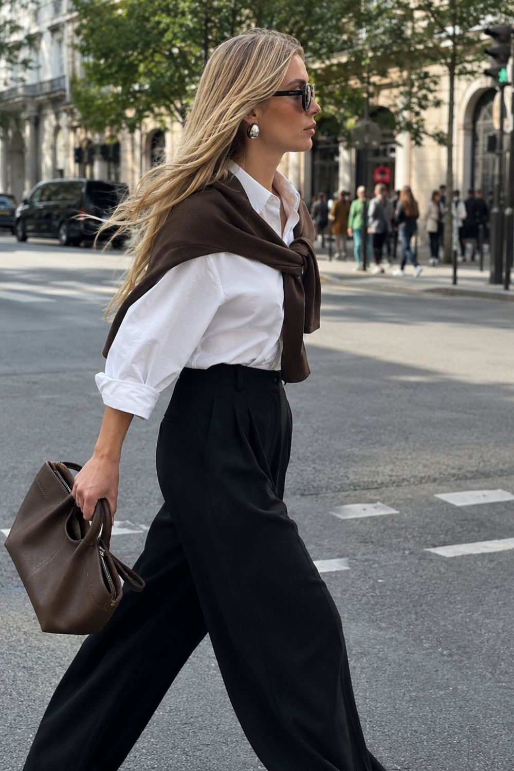 Blonde woman walking to work in a white button-down shirt, black wide-leg trousers and brown accessories, demonstrating a timeless everyday work wardrobe that fits a modern lifestyle